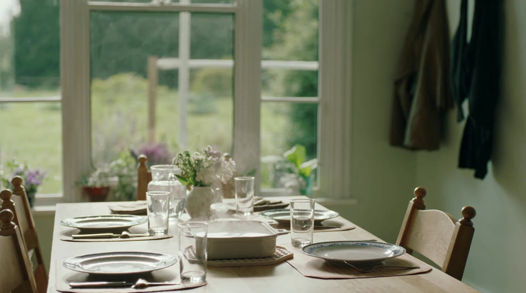 A kitchen table in an ordinary house laid for a family meal, with plates, glasses, a jug of water and a steaming casserole dish, a window onto a green garden behind and coats hanging on hooks to one side.